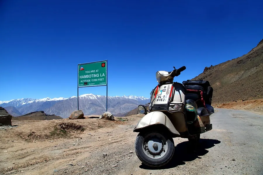 A scooter parked by the road at a high Himalayan pass with snow peaks behind